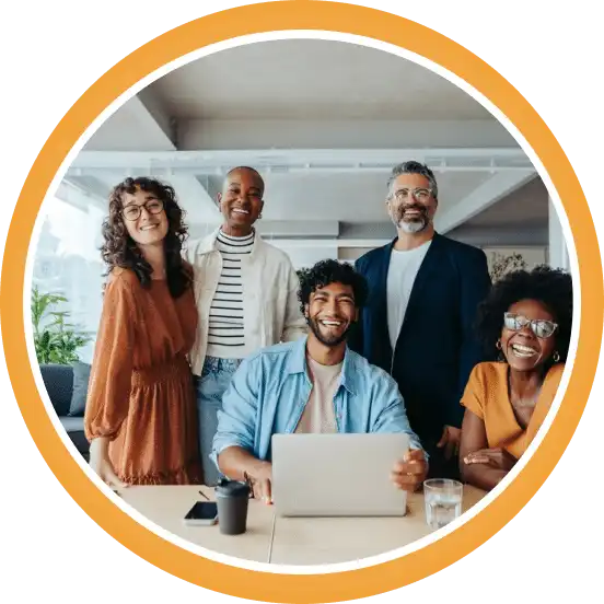A group of five people smiling at a desk; one is using a laptop. They're in a bright, modern office space with plants and large windows.